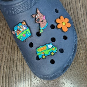 Pode incluir: Crocs azuis com pingentes de sapato temáticos do Scooby Doo. Os pingentes incluem Scooby Doo, a Mystery Machine e uma flor.