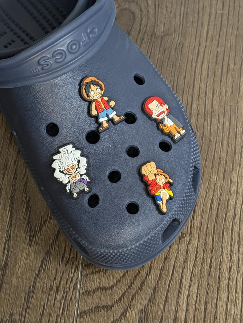 May include: Blue Crocs shoe with four colorful cartoon character shoe charms. The charms are of a red-haired character with a straw hat, a red-haired character with a beard, a white-haired character with a black robe, and a red-haired character with a straw hat.