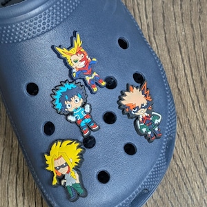 Può includere: Quattro ciondoli per scarpe colorati con personaggi dei cartoni animati che rappresentano personaggi dell'anime My Hero Academia. I ciondoli sono attaccati a un paio di zoccoli blu.