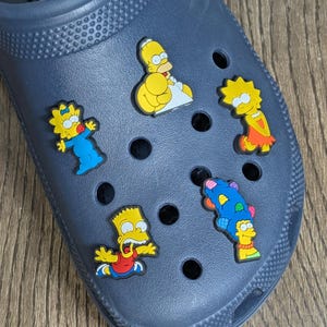 Puede incluir: Un par de Crocs azules con seis dijes de zapatos de personajes de Los Simpsons. Los dijes incluyen a Homero, Marge, Bart, Lisa, Maggie y un personaje azul con un peinado rosa y amarillo.