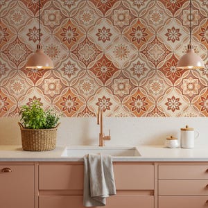 Op de afbeelding: Een keuken met een betegelde achterwand met patronen in oranje, bruin en crème. De keuken heeft een lichtroze aanrechtblad en kasten, een witte spoelbak en een koperen kraan. Een rieten mand met een plant en twee hanglampen zijn ook zichtbaar.