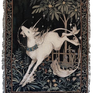 Puede incluir: Manta tejida que representa un unicornio blanco saltando frente a un árbol y una valla. El fondo es negro con un diseño floral. La manta tiene un borde con flecos.