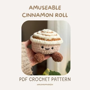 Puede incluir: Un peluche de rollo de canela tejido a ganchillo con una cara sonriente y pequeños pies marrones. El peluche tiene una base rosa claro, una cobertura blanca en la parte superior y un remolino marrón. El texto en la imagen dice "AMUSEABLE CINNAMON ROLL" y "PDF CROCHET PATTERN".