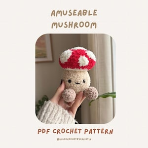 Könnte beinhalten: Ein gehäkelter Pilz mit rotem und weißem Hut, beigem Stiel und kleinen beigen Füßen. Der Pilz hat ein lächelndes Gesicht. Der Text auf dem Bild lautet "AMUSEABLE MUSHROOM" und "PDF CROCHET PATTERN".