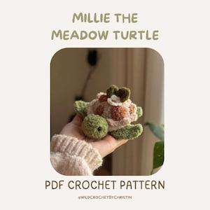 Può includere: Una tartaruga all'uncinetto fatta a mano con testa e arti verdi, guscio marrone e beige e un fiore sulla testa. Il testo "MILLIE THE MEADOW TURTLE" e "PDF CROCHET PATTERN" sono visualizzati sopra e sotto.