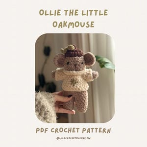 May include: A crocheted Ollie the Little Oakmouse doll, wearing a brown hat and a cream-colored sweater. The doll is held in front of a soft background. Text on the image reads "Ollie the Little Oakmouse" and "PDF Crochet Pattern".