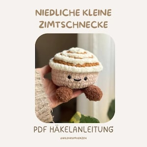 以下が含まれることがあります： ピンク、白、茶色の色合いで、笑顔の顔をしたかぎ針編みのシナモンロール。上部のテキストは「Niedliche Kleine Zimtschnecke」、下部のテキストは「PDF Häkelanleitung」と書かれています。