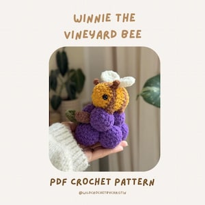May include: A handmade crochet bee with a yellow and brown head, white wings, and a purple body. The text "WINNIE THE VINEYARD BEE" and "PDF CROCHET PATTERN" are displayed above and below the bee.