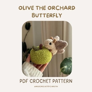 Könnte beinhalten: Ein gehäkelter Schmetterling mit hellbeigem Körper und Flügeln, der einen grünen Apfel hält. Der Text "OLIVE THE ORCHARD BUTTERFLY" und "PDF CROCHET PATTERN" sind oberhalb und unterhalb des Bildes zu sehen.