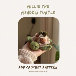 Könnte beinhalten: Eine handgefertigte Häkelschildkröte mit grünem Kopf und Gliedmaßen, einem braunen und beigefarbenen Panzer und einer Blume auf dem Kopf. Der Text "MILLIE THE MEADOW TURTLE" und "PDF CROCHET PATTERN" sind zu sehen.