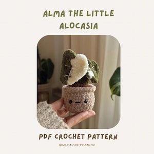 May include: A crocheted plant in a brown pot with a smiling face. The plant has green and white leaves. The text on the image says "ALMA THE LITTLE ALOCASIA" and "PDF CROCHET PATTERN".