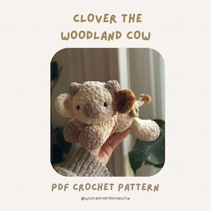 Könnte beinhalten: Gehäkeltes Kuh-Plüschtier in Creme- und Brauntönen. Das Spielzeug hat große, schlaffe Ohren und einen kleinen Schwanz. Der Text "CLOVER THE WOODLAND COW" und "PDF CROCHET PATTERN" sind oberhalb und unterhalb des Spielzeugs zu sehen.