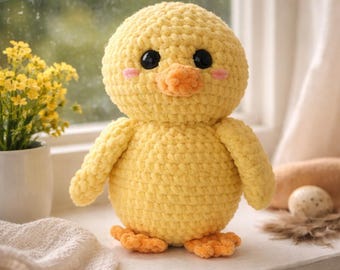 May include: A handmade, yellow crochet chick with black eyes, an orange beak, and orange feet. The chick has pink blush marks on its cheeks. The plush toy is sitting on a white windowsill, with yellow flowers in a white pot in the background.