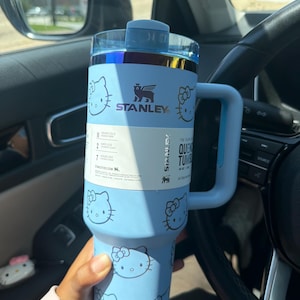 Puede incluir: Un vaso Stanley Quencher de color azul claro con una capacidad de 1,18 litros, con un patrón repetitivo de caras de Hello Kitty, un asa a juego y una tapa transparente. Una etiqueta blanca envuelve el centro mostrando las especificaciones.