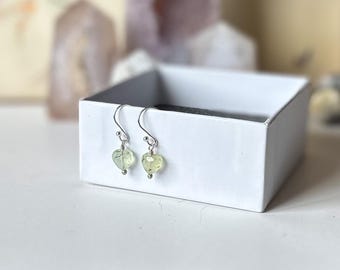 May include: A pair of silver earrings with green heart-shaped stones. The earrings are displayed in a white box.