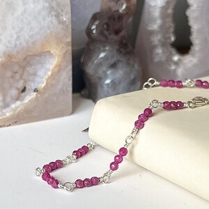 May include: A delicate silver chain bracelet with pink gemstone beads. The bracelet is on a white surface.
