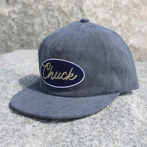 May include: A gray baseball cap with a curved brim, featuring an oval patch with the name "Chuck" in cursive script. The hat is set against a natural, rocky background.