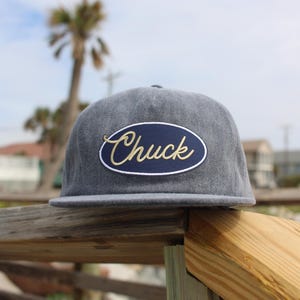 May include: A gray flat-brimmed cap with a navy blue patch featuring the name "Chuck" in gold script. The hat is resting on a wooden railing, with a blurred beach scene in the background.