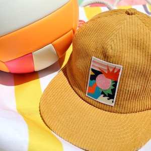 May include: Mustard yellow corduroy baseball cap with a colorful patch. The patch features a pink circle, orange sun, and abstract shapes. A striped beach towel and a colorful ball are in the background.