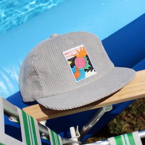 May include: A light gray corduroy baseball cap with a colorful embroidered patch featuring abstract shapes. The cap is resting on a wooden armrest of a green and white striped lawn chair, with a blue pool in the background.