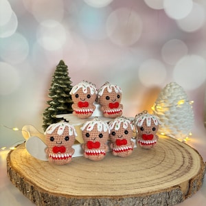 May include: A collection of handmade crochet gingerbread man keychains. Each keychain features a tan body, white icing, a red bow tie, and a smiling face. The keychains are displayed on a wooden slice with a small Christmas tree and decorative lights.