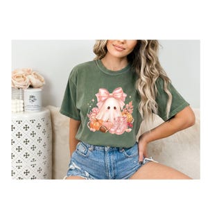 Puede incluir: Camiseta verde musgo con un diseño de fantasma con un lazo rosa, calabazas y botas. El gráfico está rodeado de follaje otoñal. La camiseta es de manga corta y de corte holgado.
