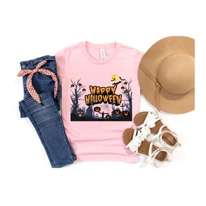 Puede incluir: Una camiseta rosa claro con las palabras "Happy Halloween" en naranja y negro, junto con un gráfico espeluznante. La imagen también incluye vaqueros azules, un sombrero beige y sandalias blancas.