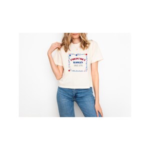 Puede incluir: Camiseta color crema con el texto "INDEPENDENT WOMAN SINCE 1776" en un marco floral rojo y azul. La camiseta se combina con unos vaqueros azules.