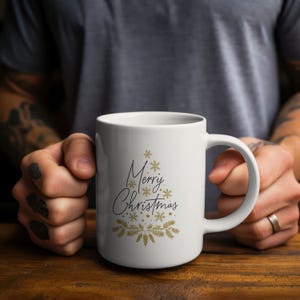 Puede incluir: Taza de cerámica blanca con un diseño de árbol de Navidad negro y dorado y la inscripción "Merry Christmas". La taza tiene un asa grande. El diseño incluye copos de nieve y ramas de pino. La taza está sujeta por una persona.