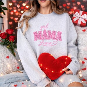 Puede incluir: Una sudadera gris claro con las palabras "girl MAMA Luna" en letras rosas. La palabra "MAMA" está rellena con un estampado floral. Se sostiene una almohada roja en forma de corazón en el regazo. Rosas y una caja de regalo en el fondo.