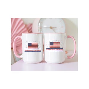 Puede incluir: Dos tazas de cerámica blancas con asas y bordes rosas. Cada taza presenta un diseño de la bandera estadounidense y el texto "MAKE AMERICA FREE AGAIN". Las tazas están sobre una superficie rosa.