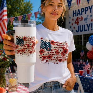 Puede incluir: Vaso y camiseta blancos con un diseño floral de la bandera estadounidense. El vaso tiene un asa y tapa negros. La camiseta y el vaso están decorados con flores rojas y blancas y estrellas azules.