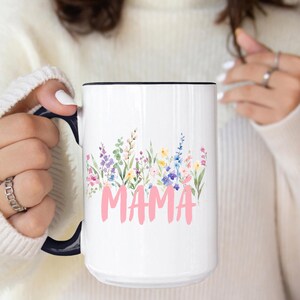 Puede incluir: Taza de cerámica blanca con asa y borde negros, con un diseño floral y la palabra "MAMA" en rosa. Las flores incluyen flores moradas, rosas, amarillas y azules. Un regalo perfecto para el Día de la Madre.