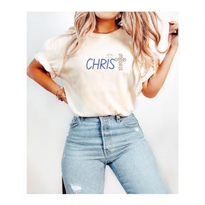 Puede incluir: Camiseta color crema con la palabra "CHRIS" en escritura azul y un diseño de cruz. La camiseta se combina con vaqueros de mezclilla azul claro. El estilo es casual y cómodo.