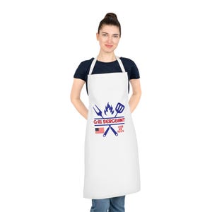 Puede incluir: Delantal blanco con un diseño patriótico. Presenta el texto "Grill Sergeant", espátula y tenedor cruzados, una llama, la bandera estadounidense y el texto "4th of July".