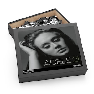 Puede incluir: Una foto en blanco y negro de Adele sobre un fondo gris. El texto "ADELE 21" está en letras blancas y verdes en la parte inferior de la imagen. La caja es para un rompecabezas de 120 piezas de 8x10 para mayores de 9 años.
