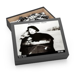 May include: A black and white 120 piece jigsaw puzzle featuring a photo of Joni Mitchell on the album cover for her album "Hejira". The album cover features Joni Mitchell wearing a black beret and a fur coat, with a road and a cloudy sky in the background.