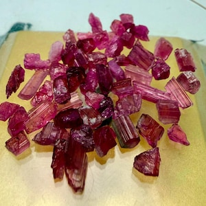 Könnte beinhalten: Eine Ansammlung von rohen, ungeschliffenen rosa und tiefroten Turmalinkristallen. Die Steine variieren in Größe und Form und weisen natürliche Streifen sowie durchscheinende bis undurchsichtige Texturen auf, die lose auf einer Fläche liegen.