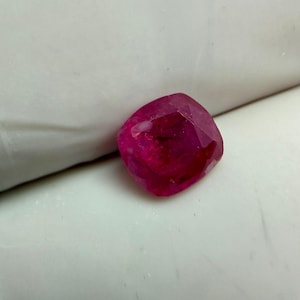 Peut inclure: Une pierre précieuse rubis taillée en coussin. La gemme est d'un rouge profond et saturé, avec de multiples facettes captant la lumière. Le rubis est posé sur un fond blanc.