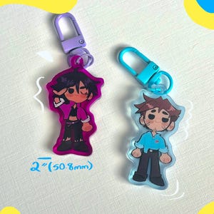 Puede incluir: Dos llaveros acr&iacute;licos con personajes de dibujos animados. Un llavero tiene un borde y un cierre morados, el otro tiene un cierre y un borde azules. Los llaveros miden aproximadamente 5 cm de alto.