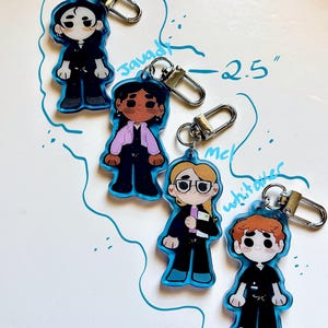 May include: Four cartoon character keychains with blue outlines and silver clasps. Each keychain is approximately 2.5 inches tall. The characters are dressed in various outfits, including a black suit, a pink jacket, and a black uniform. The names "Santos", "Javadi", "Mc", and "Whitaker" are written above or near each character.