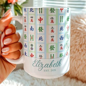 Puede incluir: Taza de cerámica blanca con un colorido diseño de fichas de Mahjong. Personalizada con el nombre "Elizabeth" y el año "EST. 2026". La taza tiene un asa blanca y es sostenida por una persona.