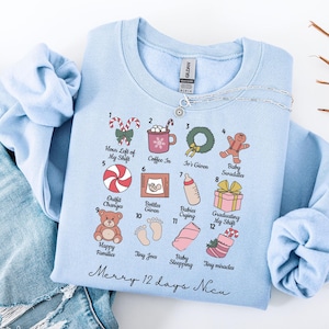 May include: A light blue crew neck sweatshirt with a graphic of 12 Christmas-themed illustrations and text. The illustrations include a candy cane, coffee cup, wreath, gingerbread man, and baby-related items. The text reads "Merry 12 days Nicu".