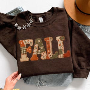 May include: A dark brown long-sleeve sweatshirt featuring the word FALL in large block letters. Each letter is filled with a patchwork pattern of floral, plaid, and striped designs in shades of orange, green, cream, and brown.