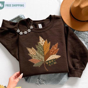 May include: A dark brown long-sleeve sweatshirt featuring a large, central maple leaf graphic composed of various patchwork-style fabric patterns in shades of green, orange, brown, and cream, accented with a silver-toned floral necklace.