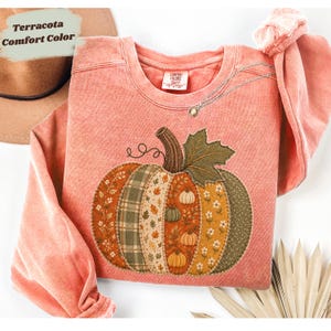 May include: A terracotta-coloured crewneck sweatshirt featuring a large, central pumpkin graphic. The pumpkin is composed of various fabric-like patterns in shades of orange, green, and cream, including floral, plaid, and leaf motifs. A silver necklace rests on the neckline.
