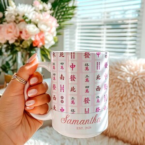 Pu&ograve; includere: Tazza in ceramica bianca con un design rosa e bianco con caratteri cinesi. La tazza &egrave; personalizzata con il nome "Samantha" e il testo "EST. 2026" in rosa.