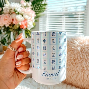 Pu&ograve; includere: Tazza in ceramica bianca con un design sfumato blu chiaro con caratteri cinesi. La tazza &egrave; personalizzata con il nome "Daniel" e l'anno di fondazione "EST. 2026" in un carattere corsivo. La tazza &egrave; tenuta in una mano.