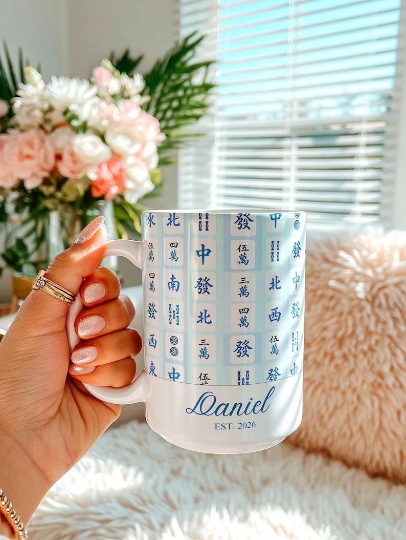 Pu&ograve; includere: Tazza in ceramica bianca con un design sfumato blu chiaro con caratteri cinesi. La tazza &egrave; personalizzata con il nome "Daniel" e l'anno di fondazione "EST. 2026" in un carattere corsivo. La tazza &egrave; tenuta in una mano.