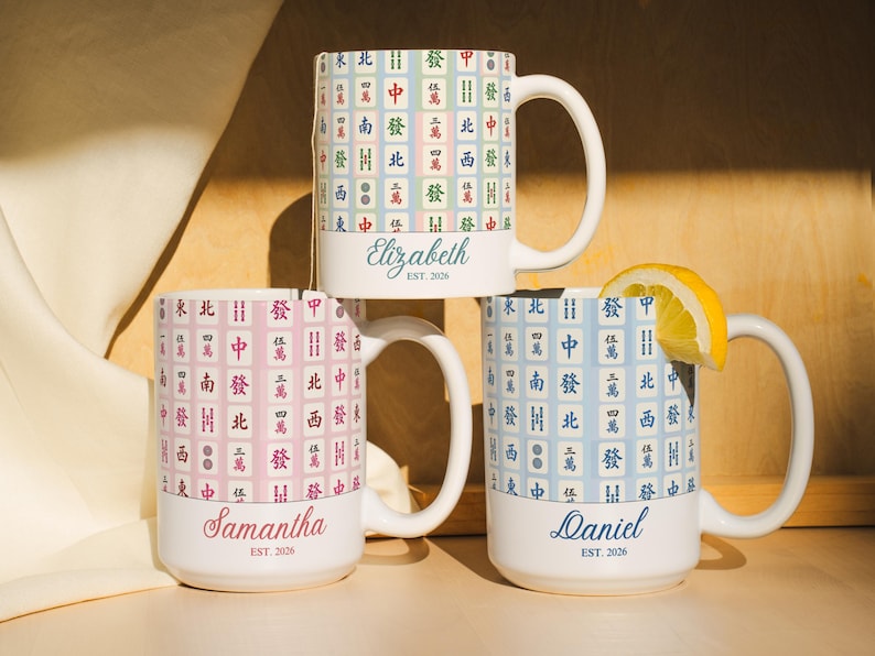 Pu&ograve; includere: Tre tazze di ceramica bianca impilate, ciascuna con uno sfondo pastello diverso e un motivo a piastrelle di Mahjong. Le tazze sono personalizzate con nomi e l'anno 2026. Una tazza ha una fetta di limone.